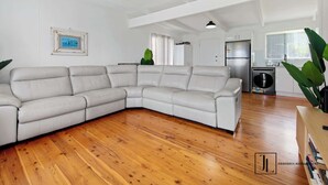 Ein geräumiger Wohnbereich mit einem großen, L-förmigen Ecksofa, Holzböden, einer Küche mit Edelstahlgeräten und Blick ins Freie.