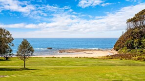 Escape to a serene coastal haven with a lush grassy area leading to a sandy beach, surrounded by rocky outcrops and lush vegetation.