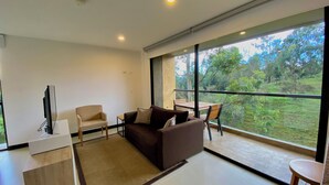 Un salon moderne avec un canapé, un téléviseur et une table à manger avec des chaises. Une grande fenêtre offre une vue sur la verdure environnante.