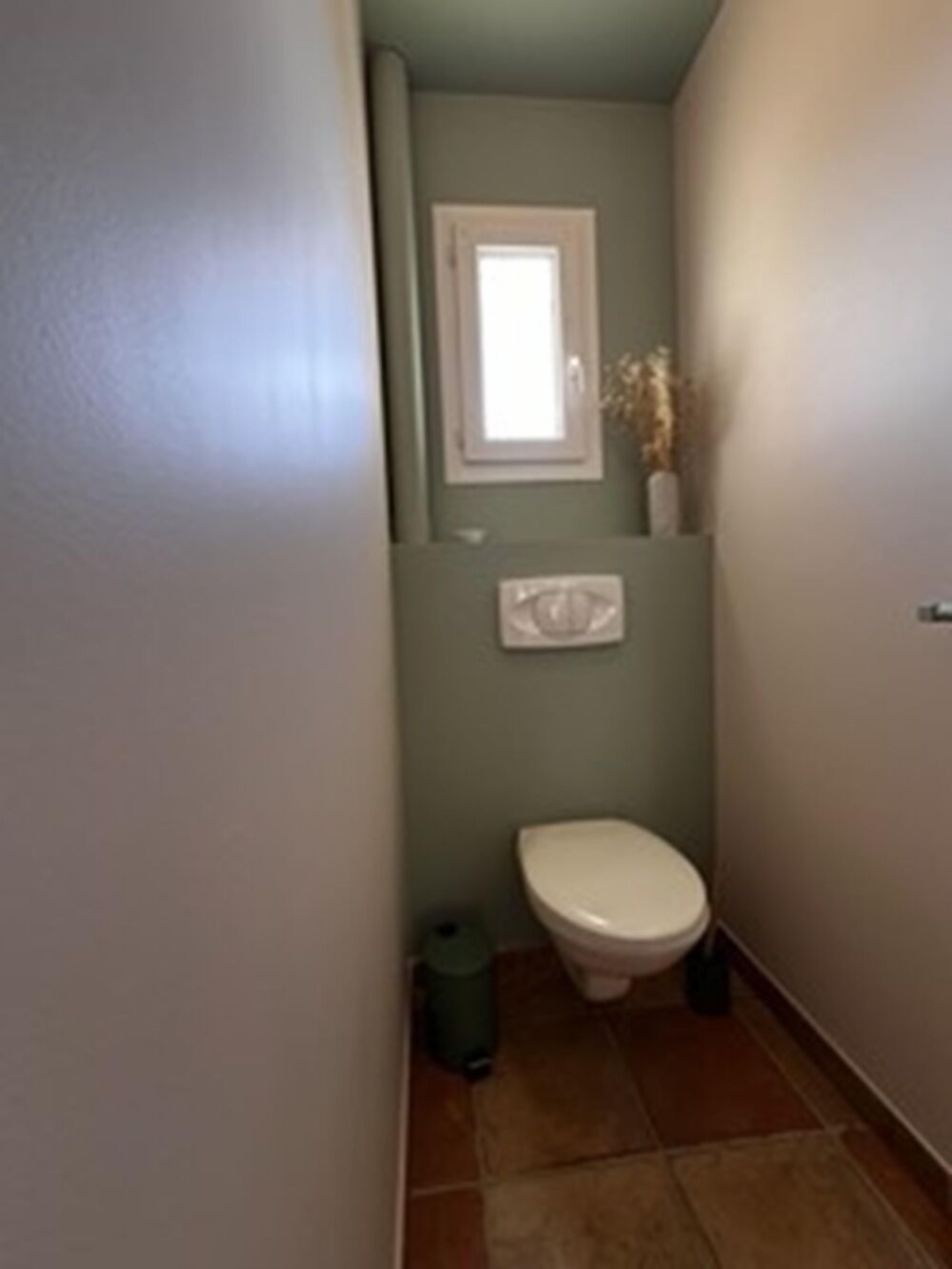 A small bathroom with a toilet, a window, and a trash can.