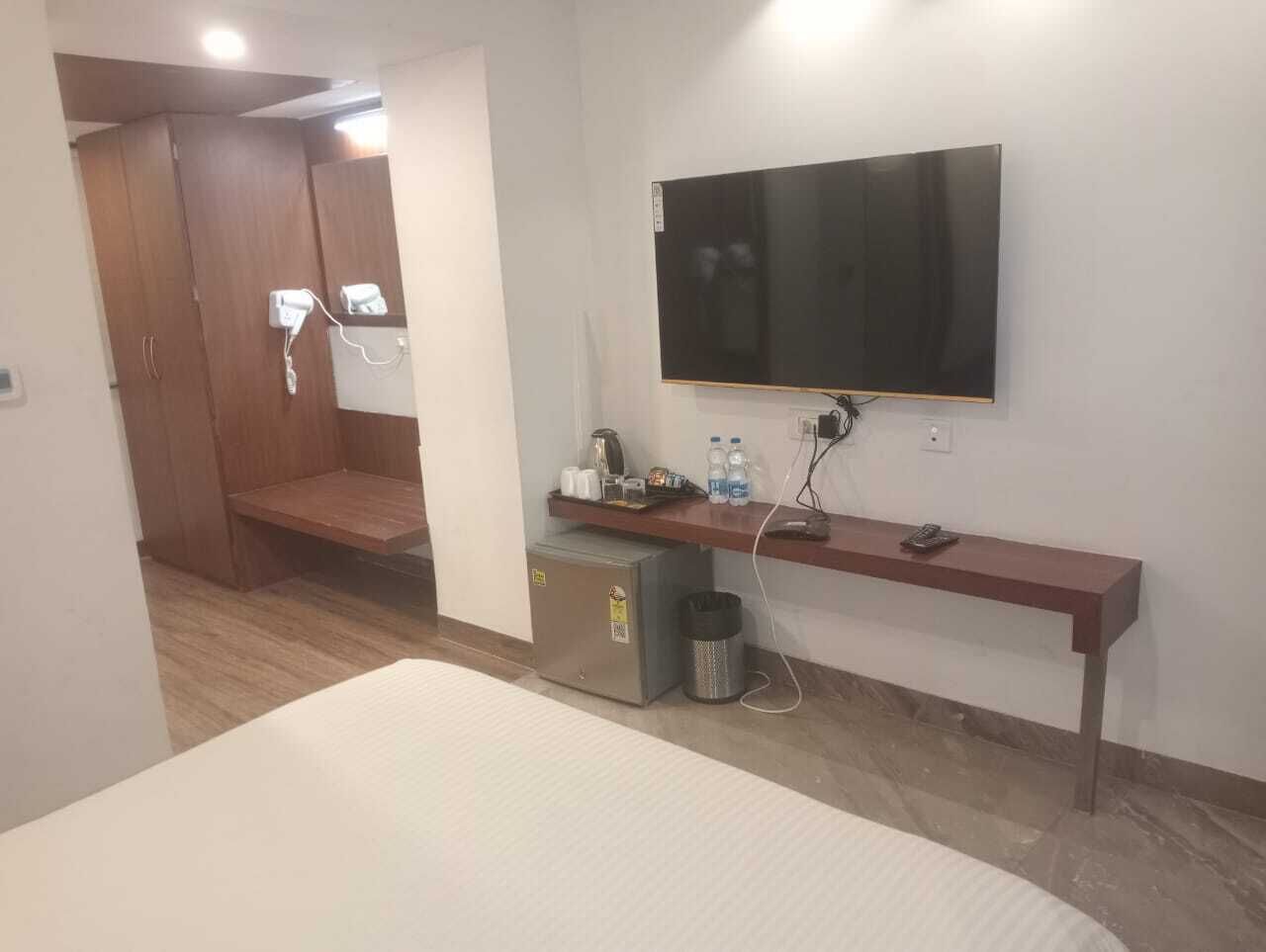 A hotel room with a bed, a TV mounted on the wall, a small desk with a chair, a trash bin, and a wooden wardrobe.