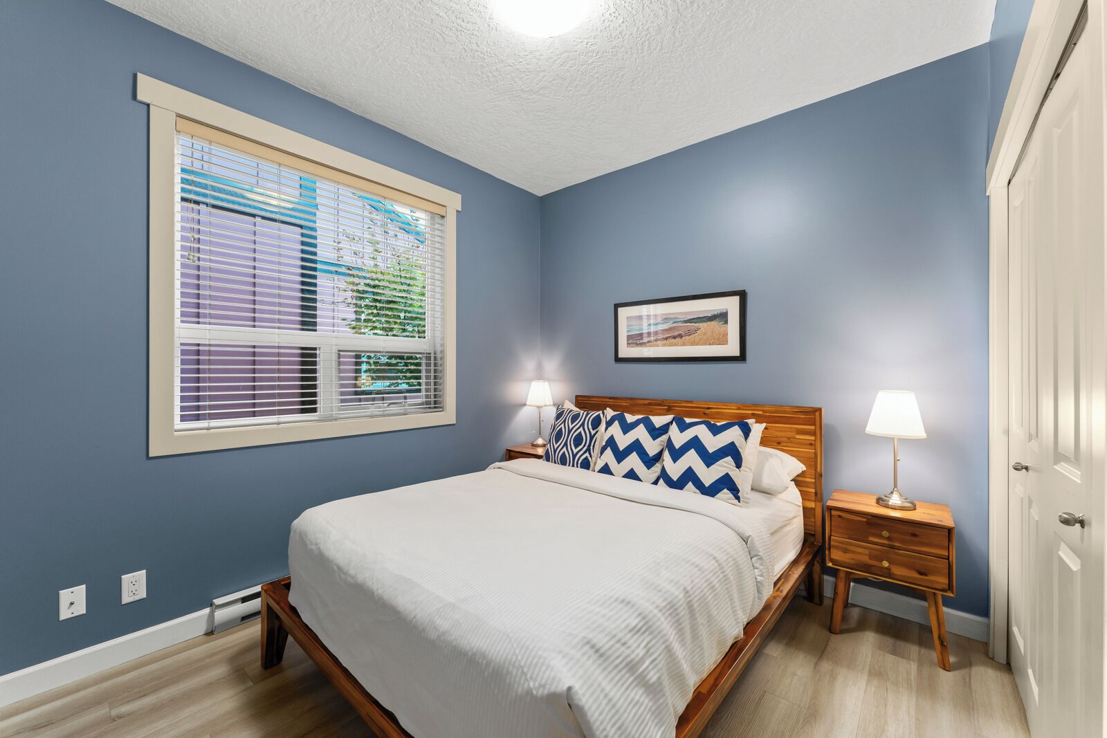 Una camera da letto con un letto, due comodini, una finestra con tende e un quadro appeso al muro, più una porta.