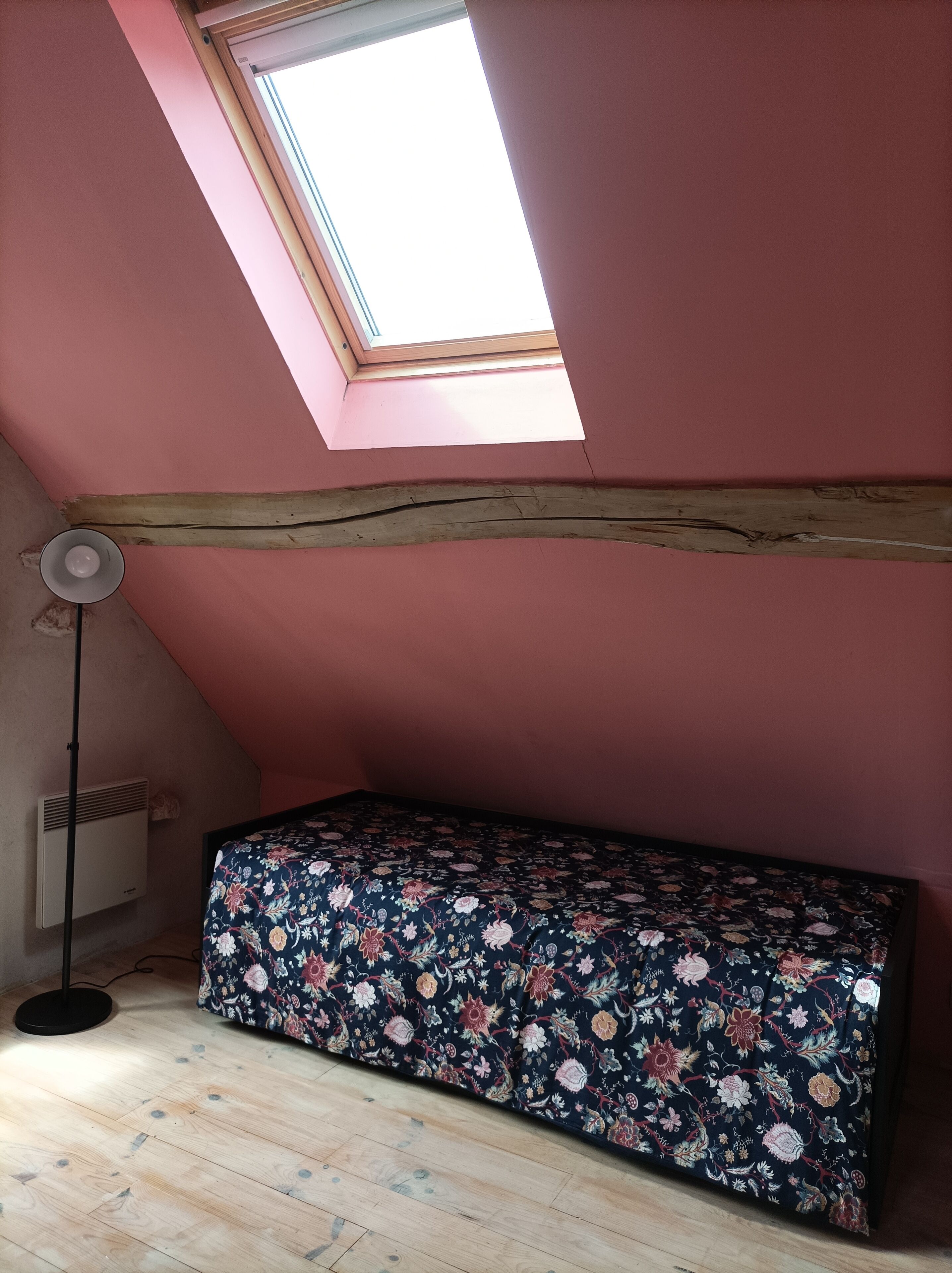 Une petite pièce agréable, avec un plafond en pente, un puits de lumière, un lit recouvert d’une couvre-lit à motifs floraux, un lampadaire et un radiateur.
