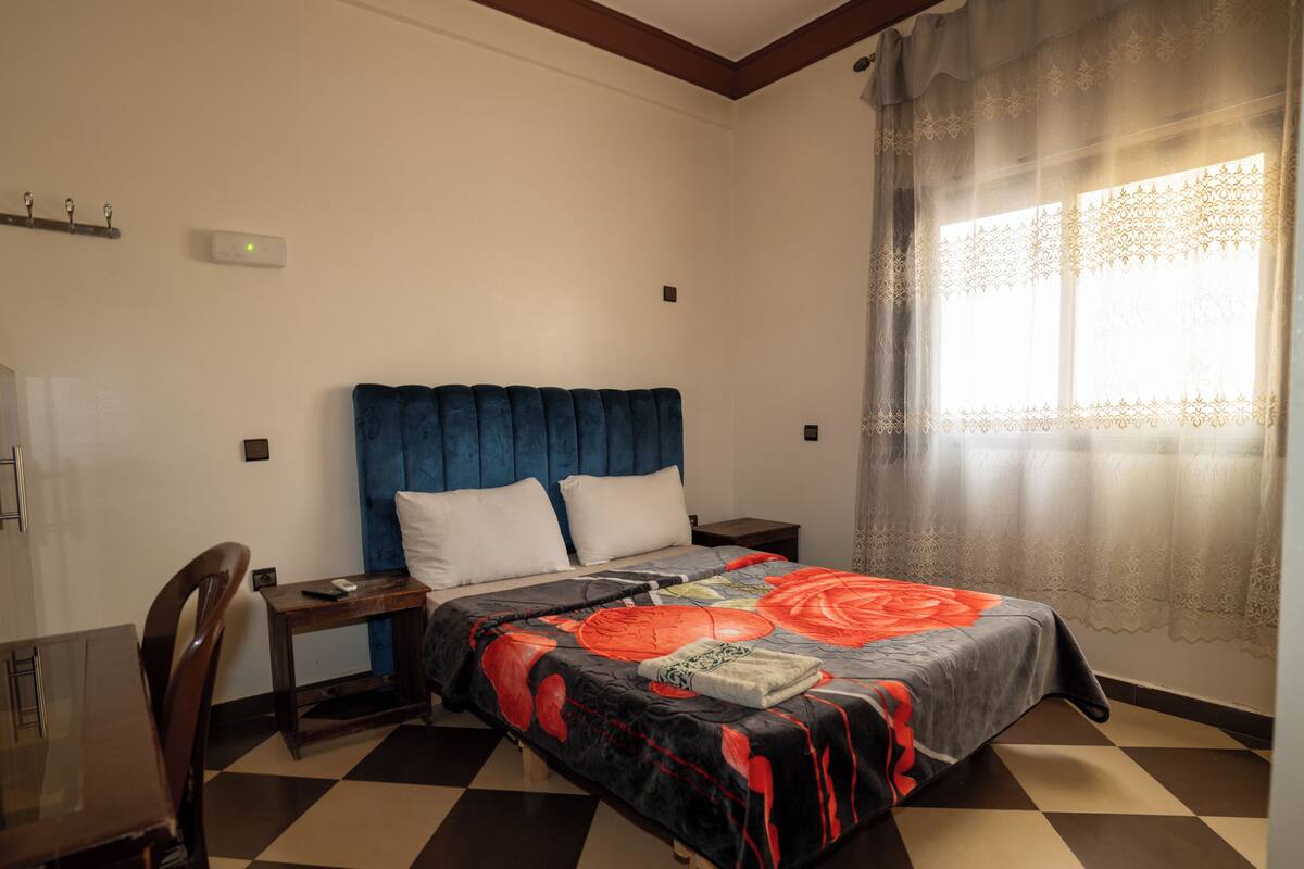 Ein Schlafzimmer mit Schachbrettmuster-Boden, einem Bett mit floralem Überwurf, einem hölzernen Nachttisch, einem Schreibtisch und einem Stuhl.