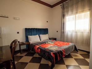 A bedroom with a checkered floor, a bed with a floral bedspread, a wooden nightstand, a desk, and a chair. - ZELLAKA hôtel & café (Khouribga)