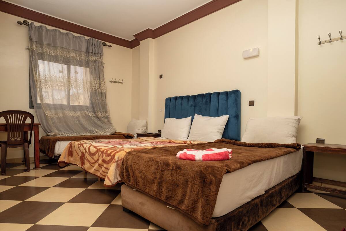 Ein Hotelzimmer mit Schachbrettboden, einem Bett mit brauner Kopfteil, einem Sessel und einem Fenster mit durchsichtigen Vorhängen.