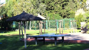 Una mesa de tenis en un parque con un pabellón de techo de paja y una valla verde.