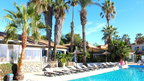 A pool area with lounge chairs, palm trees, and a stone building. - Malibu Village (Canet-en-Roussillon)