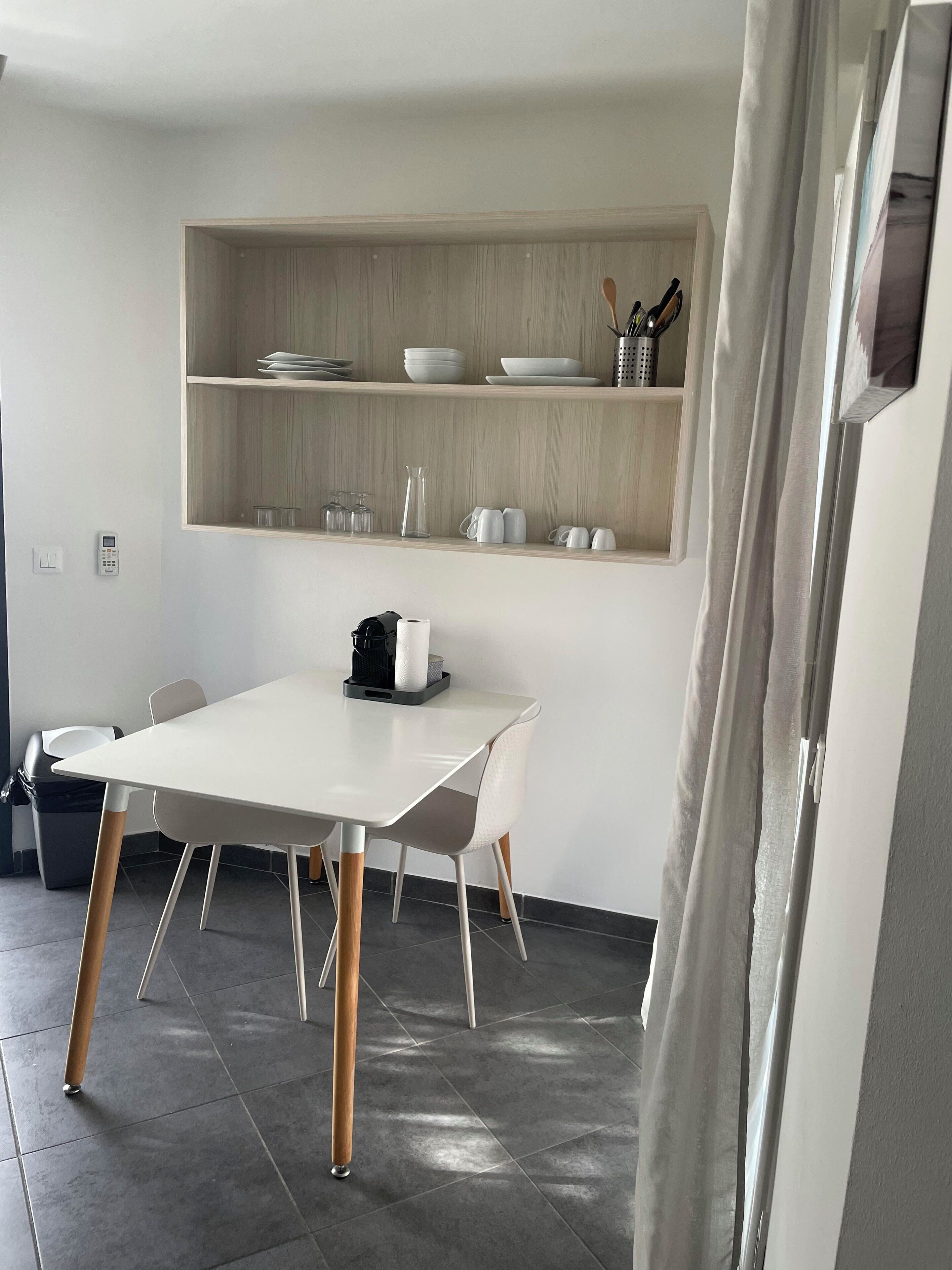 A modern kitchen with a white table, two chairs, and a coffee machine. There are shelves above the counter holding dishes and utensils.
