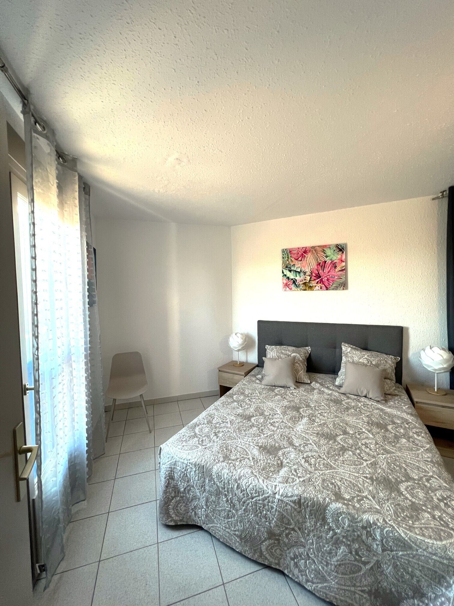 A bedroom with a large bed, a nightstand, a chair, a window with curtains, and a floral artwork on the wall.