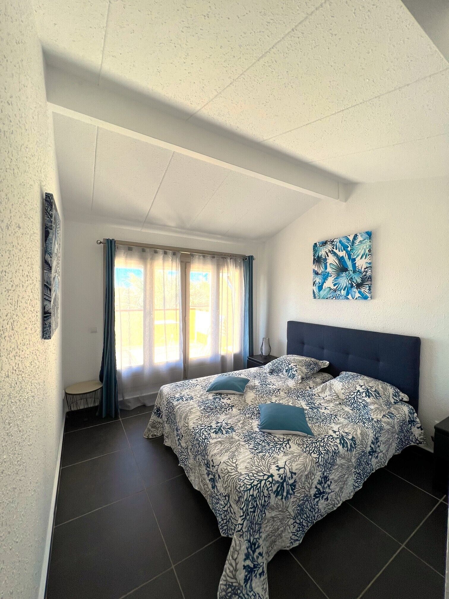 A bedroom with a bed, a window with curtains, a painting on the wall, and a small table.