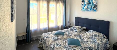 A bedroom with a bed, a window with curtains, a painting on the wall, and a small table.