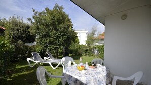 Een terras met een tafel gedekt voor twee personen, omringd door witte stoelen, een kleine tafel met een fruitschaal en twee ligbedden.