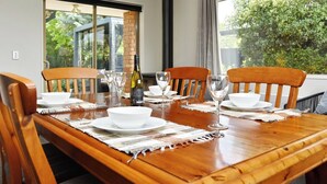 A dining table set for four with white plates, bowls, and wine glasses, in a room with large windows and a view of greenery outside. - Stay at Puhara - Parklands Escape (Christchurch)