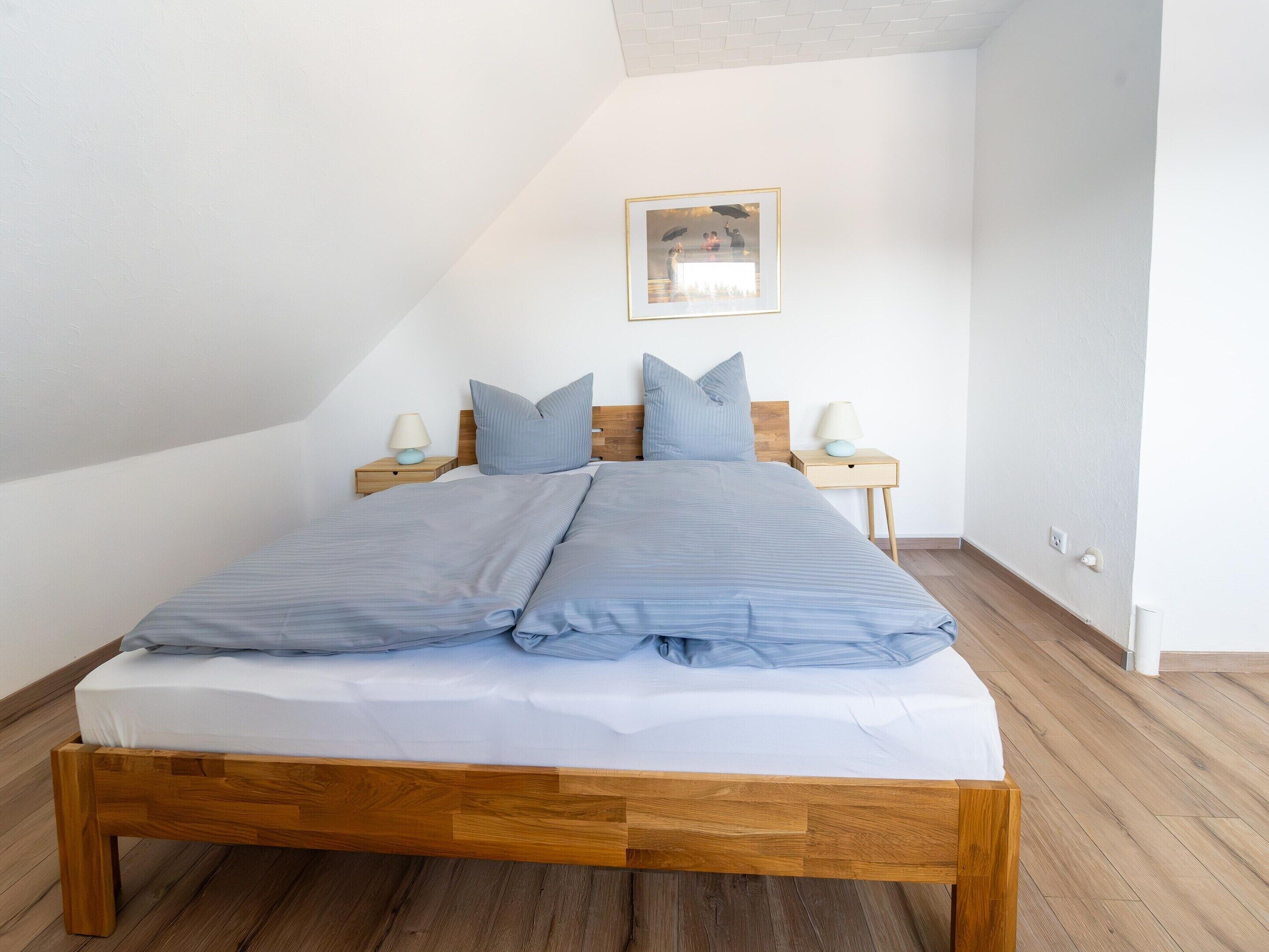 A neatly made bed with a wooden frame and blue bedding, two bedside tables with lamps, and a framed picture on the wall.