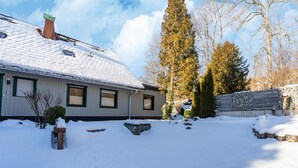 A snow-covered house with a chimney, a stone wall, and a tall evergreen tree. - Forest Villa With Sauna (Oberharz am Brocken)