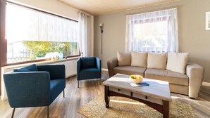 A living room with a sofa, two armchairs, a coffee table, and a view of a garden through large windows. - Forest Villa With Sauna (Oberharz am Brocken)