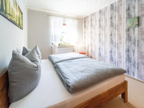 A bedroom with a wooden bed, grey pillows, a white nightstand, and a window with sheer curtains.