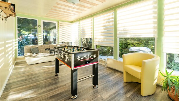 A cozy living room with a foosball table, a sofa, a yellow armchair, and large windows with blinds. - Forest Villa With Sauna (Oberharz am Brocken)