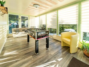 A cozy living room with a foosball table, a sofa, a yellow armchair, and large windows with blinds. - Forest Villa With Sauna (Oberharz am Brocken)