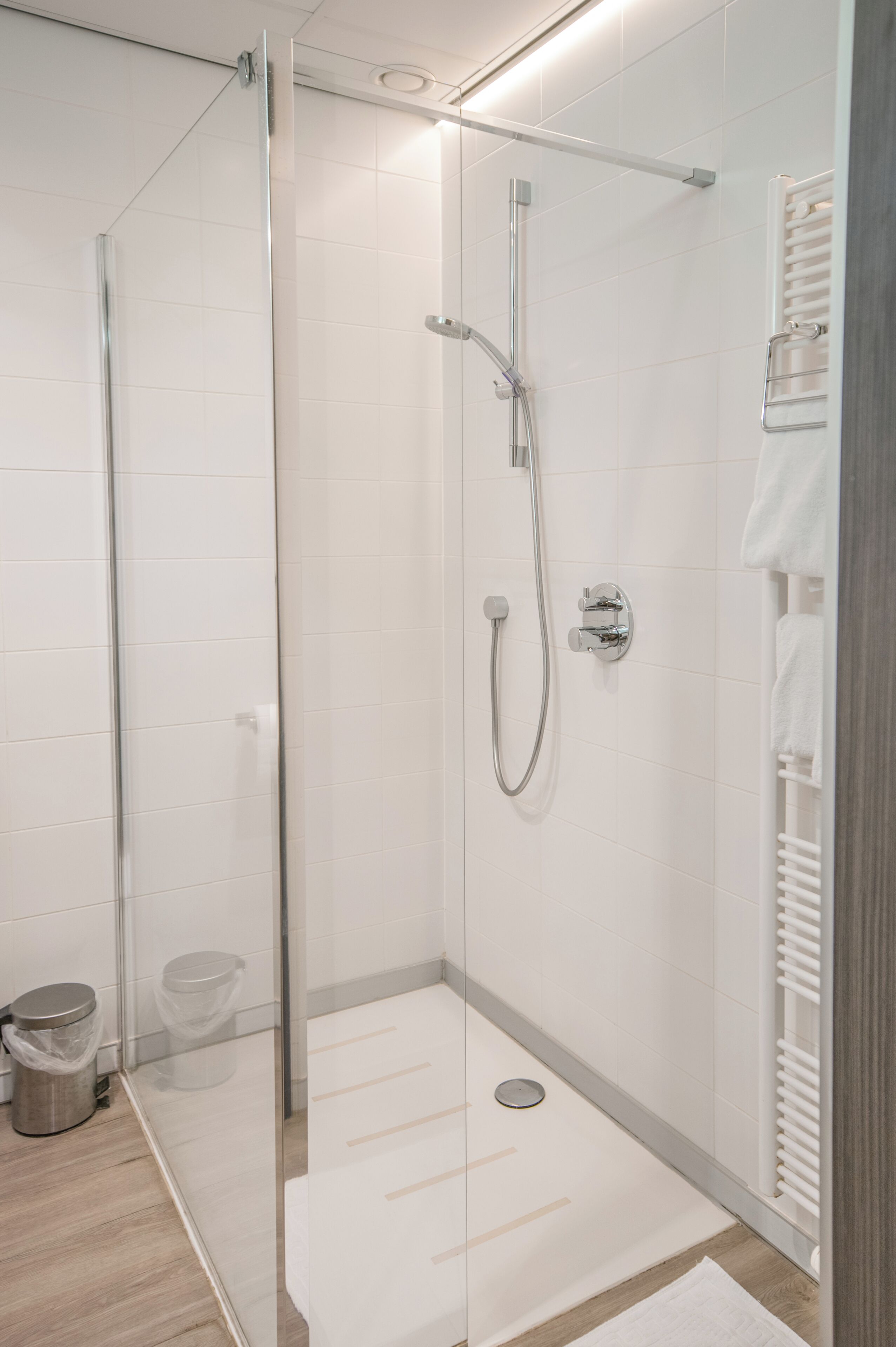 Un baño moderno con ducha envuelta en cristal, pared de azulejos blancos, toallero blanco y suelo de madera.