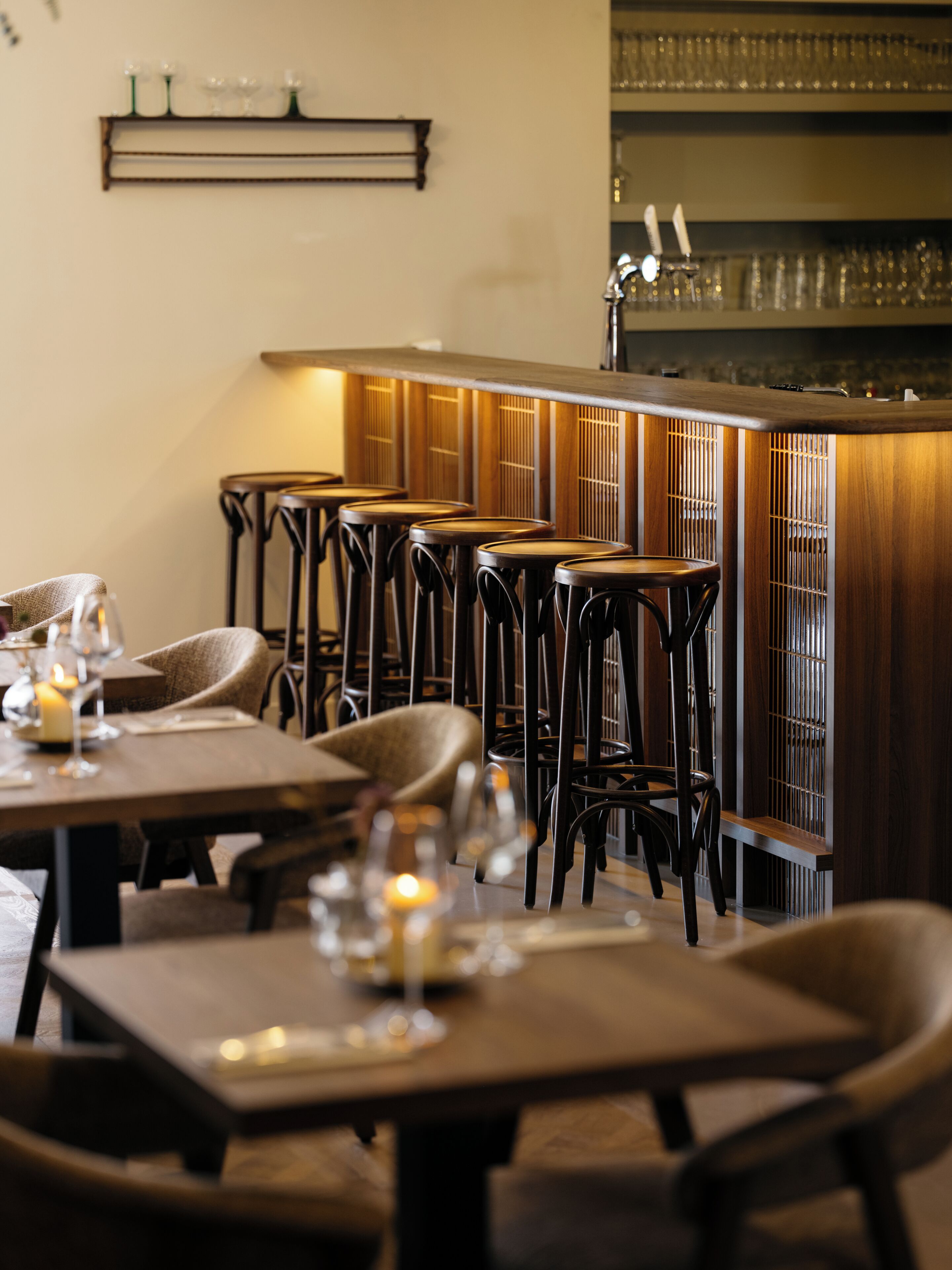 A bar area with wooden stools and tables, a bar counter, and shelves with glasses.