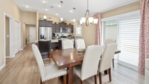 Un comedor con mesa de madera y sillas blancas tapizadas, una cocina al fondo y un ventanal con persianas.