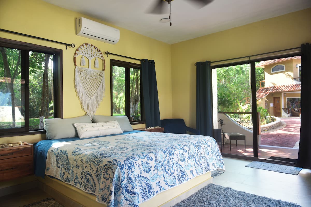 Une chambre à coucher avec un lit, une décoration murale en macramé, une fenêtre et une vue sur l’extérieur.