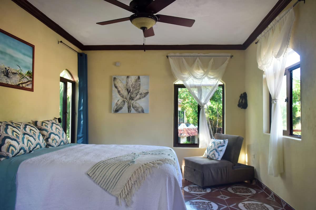 Une chambre à coucher avec un lit, un ventilateur de plafond, une fenêtre avec des rideaux, un tableau et un canapé.