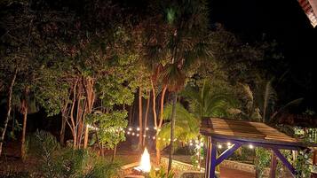 An outdoor seating area with a fire pit, surrounded by lush greenery and a wooden pergola.
