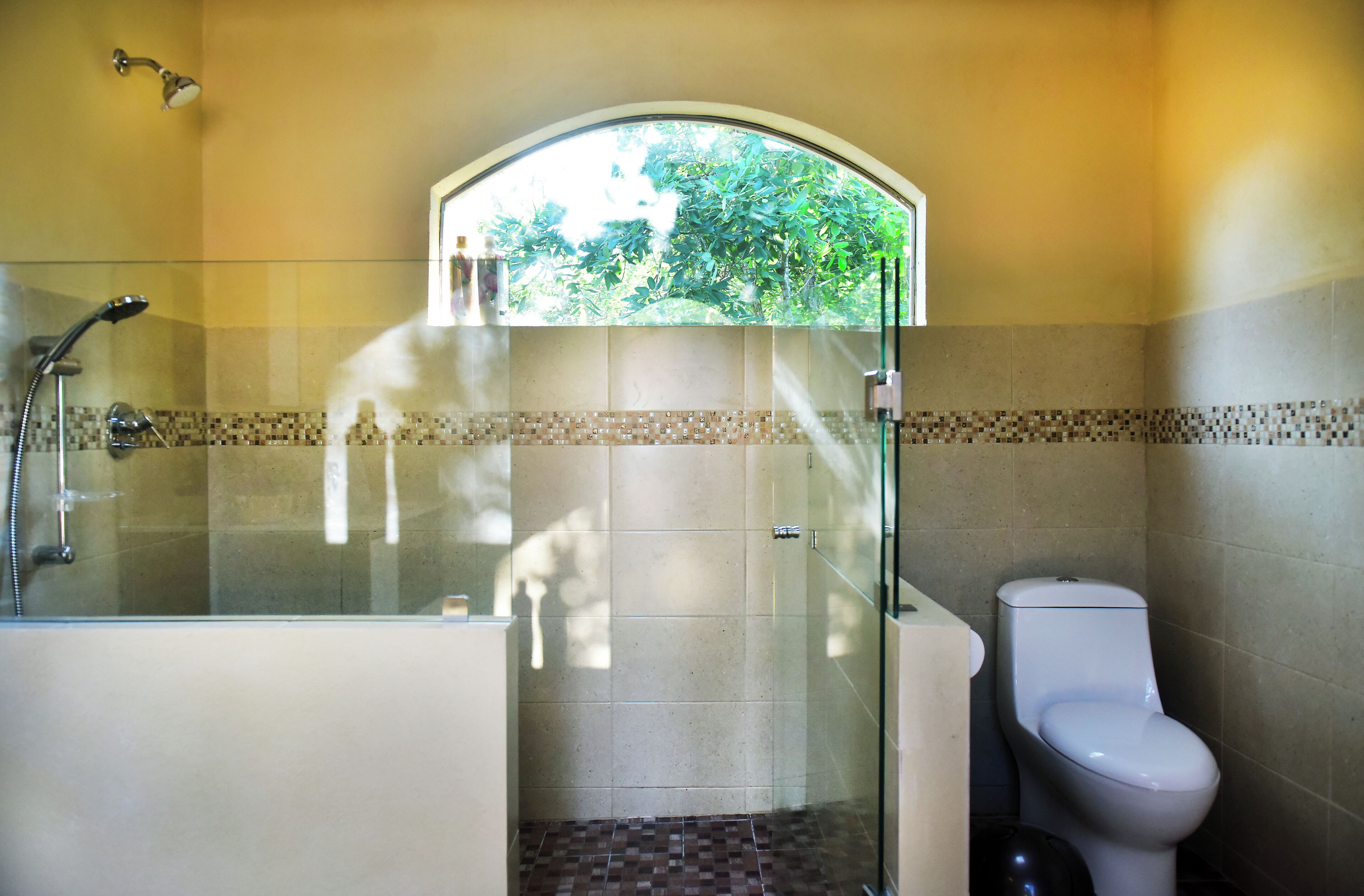 A bathroom with a glass-enclosed shower, a toilet, and a window with a view of greenery.