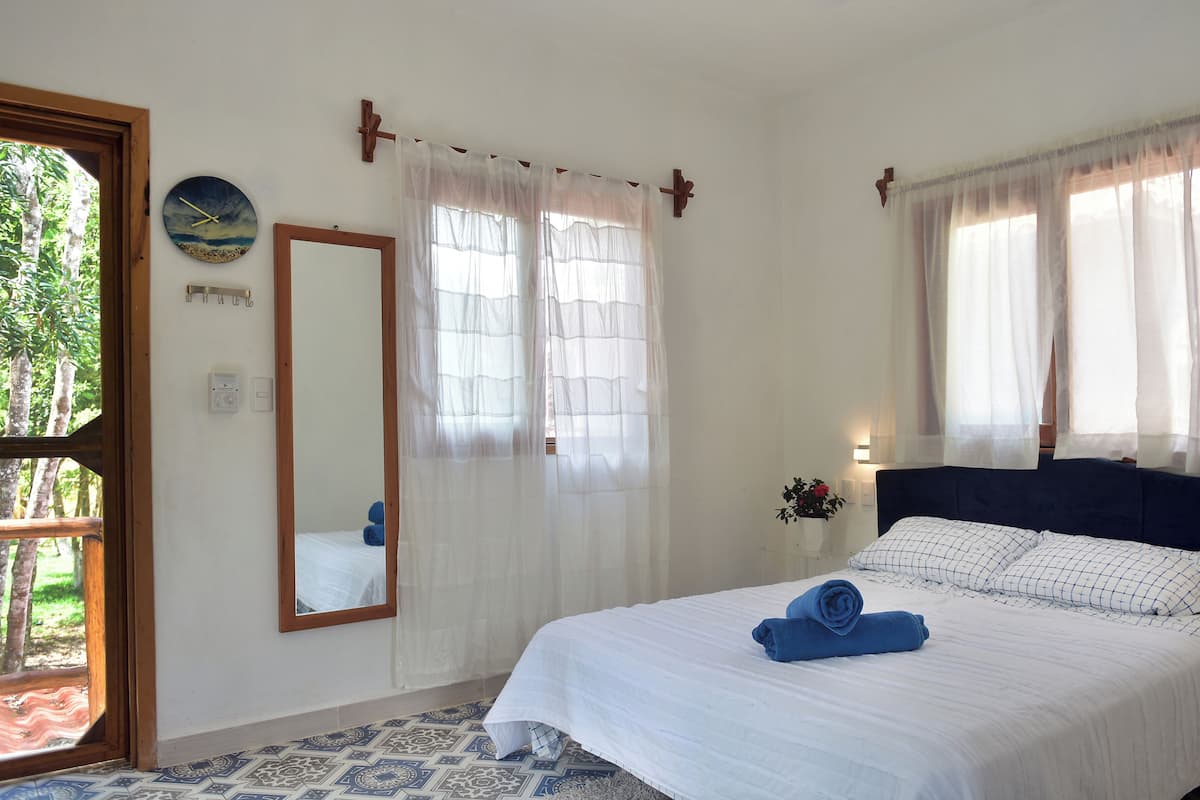Une chambre à coucher avec un lit, un miroir, une fenêtre avec des rideaux légers et une vue sur les arbres à l’extérieur.