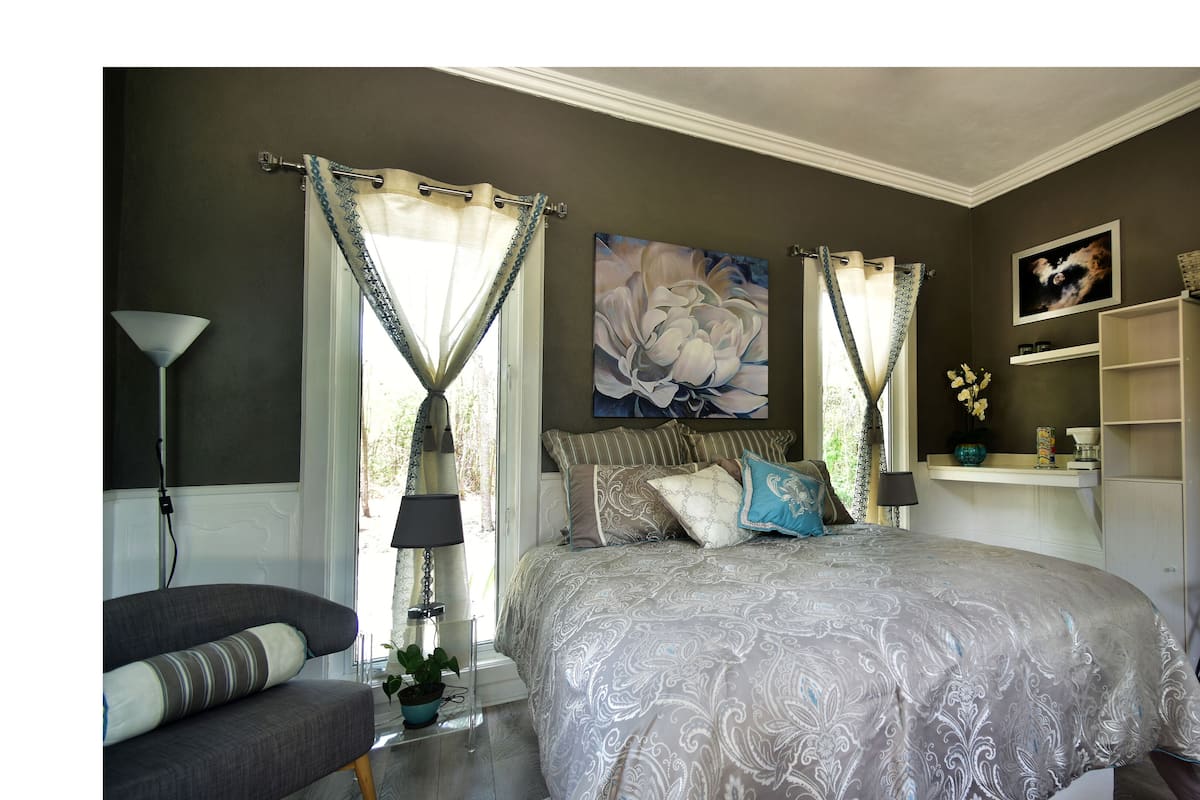 Une chambre à coucher avec un grand lit, une table de chevet, une chaise, une lampe, une fenêtre avec des rideaux et une étagère fixée au mur, ornée d’objets décoratifs.