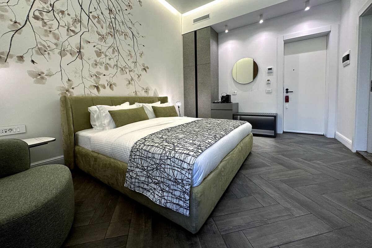 A modern bedroom with a large bed, a bedside table, a chair, a wall-mounted mirror, and a door.