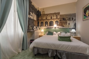 Quarto de hotel com uma cama grande, mesa de cabeceira com abajur, espelho fixado na parede e um mural de um edifício antigo.
