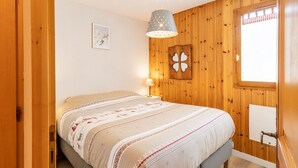 A cozy bedroom with a wooden wall panel, a bed with a patterned bedspread, a bedside lamp, and a window with a checkered curtain.