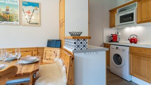 A compact kitchen with wooden cabinets, a washing machine, a microwave, and a dining area with a table set for four.
