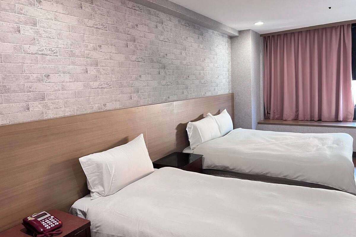 Habitación de hotel con dos camas, una pared de ladrillo, un cabecero de madera, un teléfono y una ventana con cortinas.