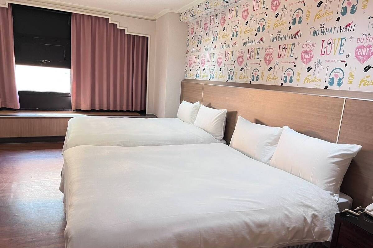 Habitación de hotel con dos camas, un ventanal grande con cortinas y una pared decorada con diversas palabras y frases.
