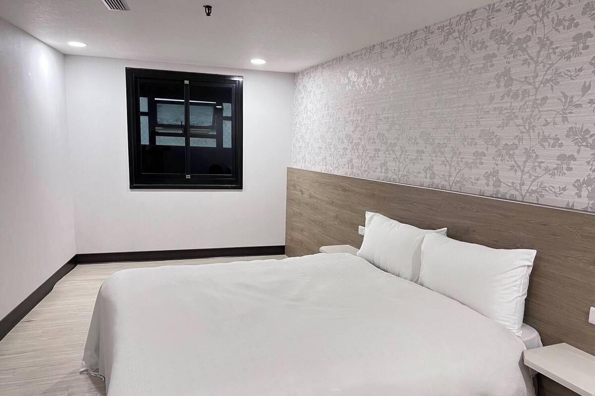 Una cama impecable con ropa blanca en una habitación con cabecera de madera, un ventanal pequeño y un papel tapiz con motivos florales.