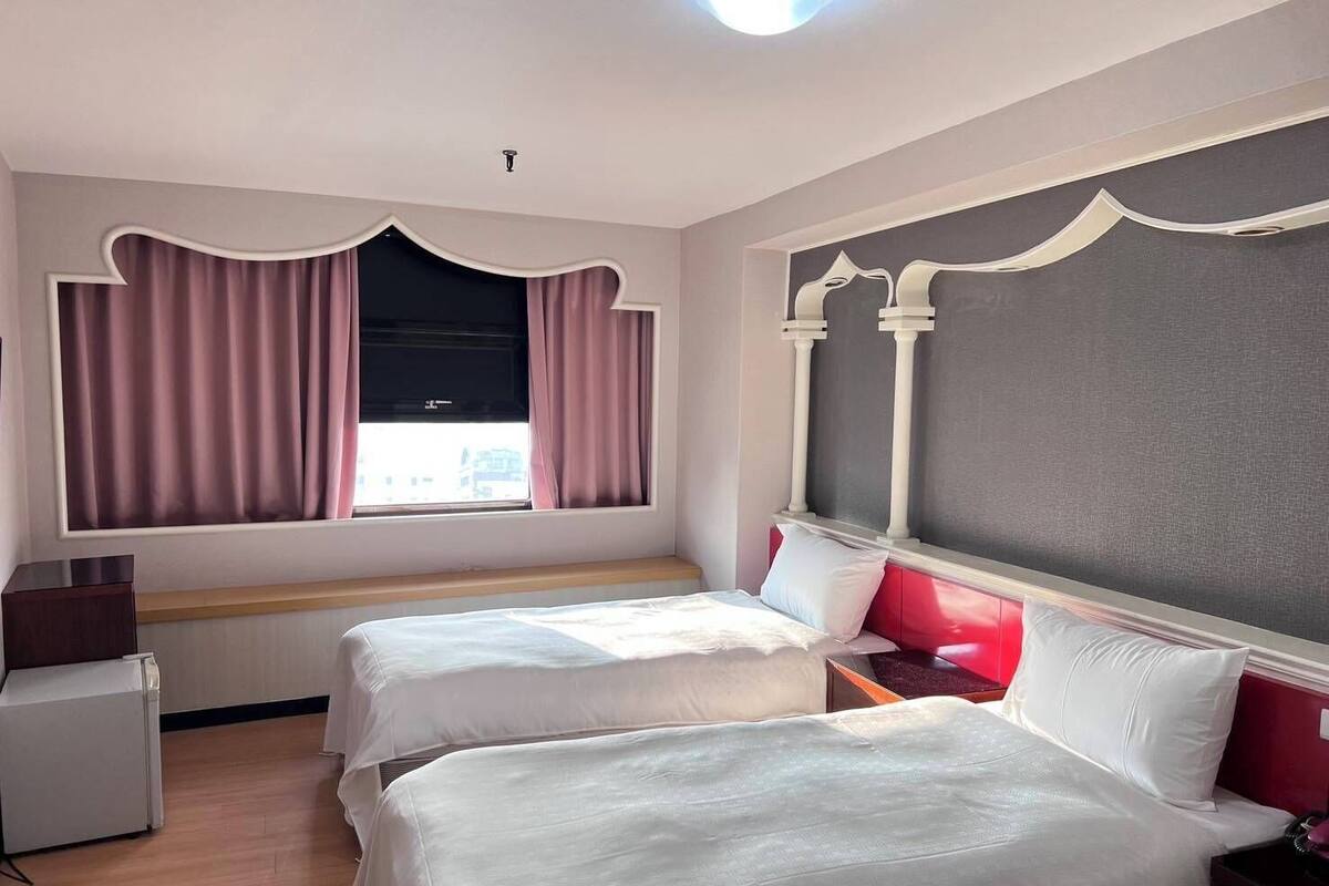 Habitación de hotel con dos camas, una ventana con cortinas, un radiador y un pequeño escritorio con lámpara.