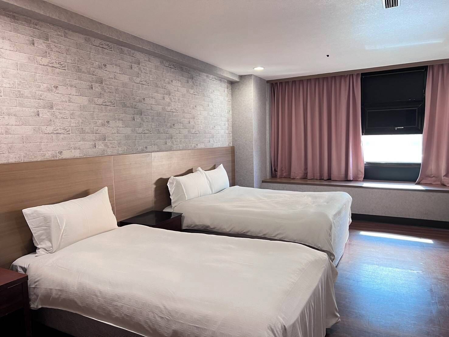 A hotel room with two beds, a brick wall, a window with curtains, and a wooden headboard.