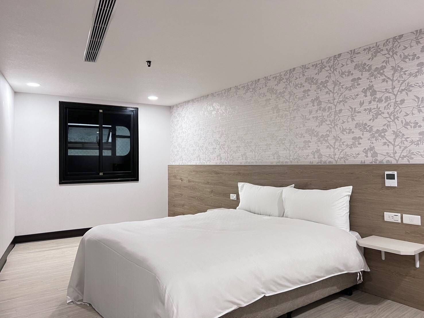 A modern hotel room with a large bed, a wooden headboard, a bedside table, and a window with blinds.