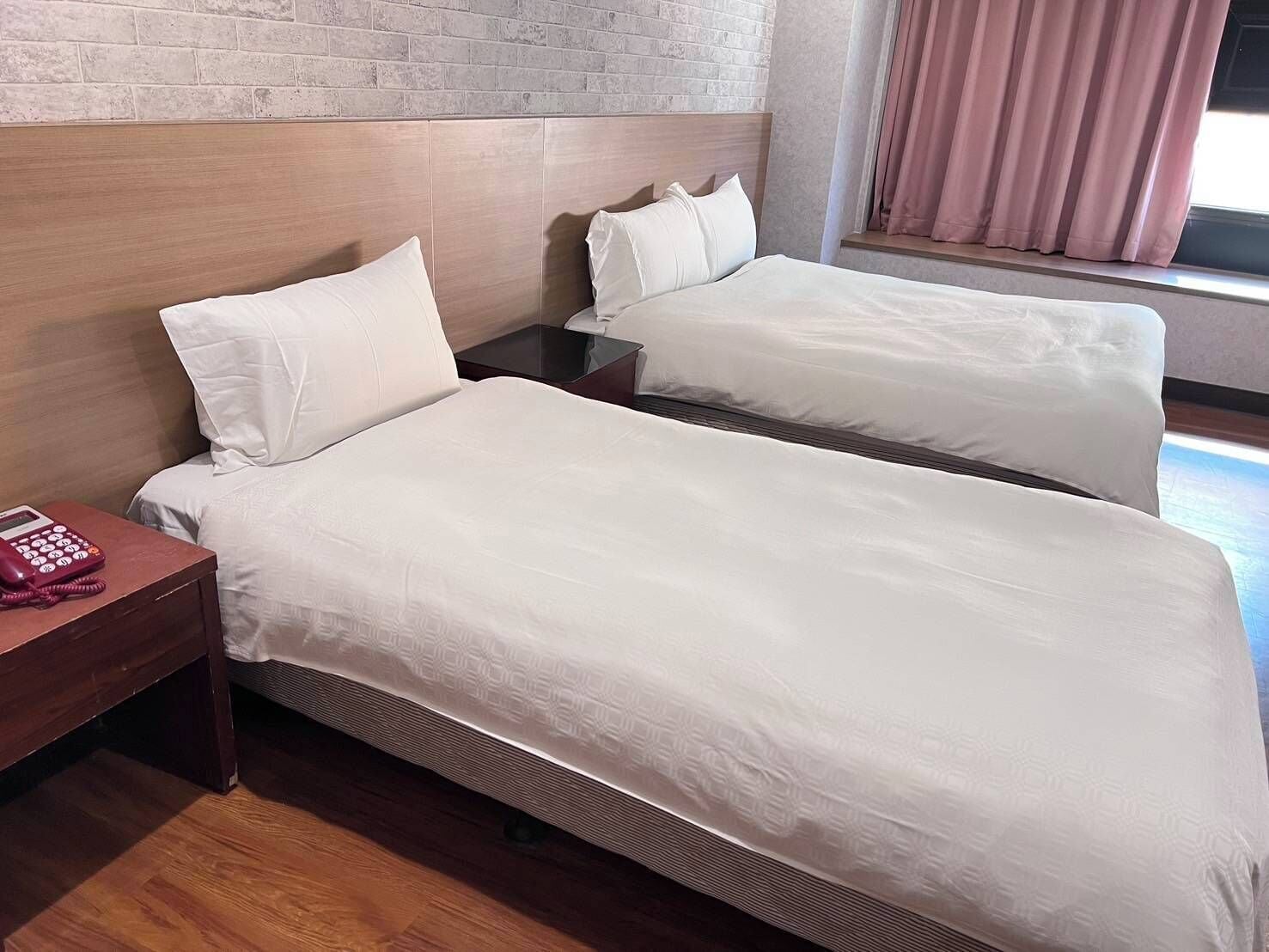 Une chambre d’hôtel avec deux lits, une tête de lit en bois, une table de chevet avec un téléphone et une fenêtre avec des rideaux.