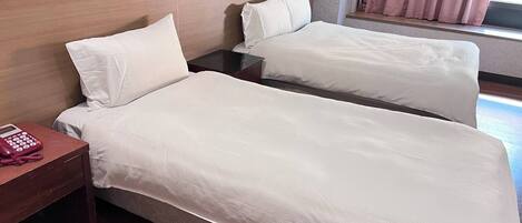 A hotel room with two beds, a wooden headboard, a bedside table with a phone, and a window with curtains.