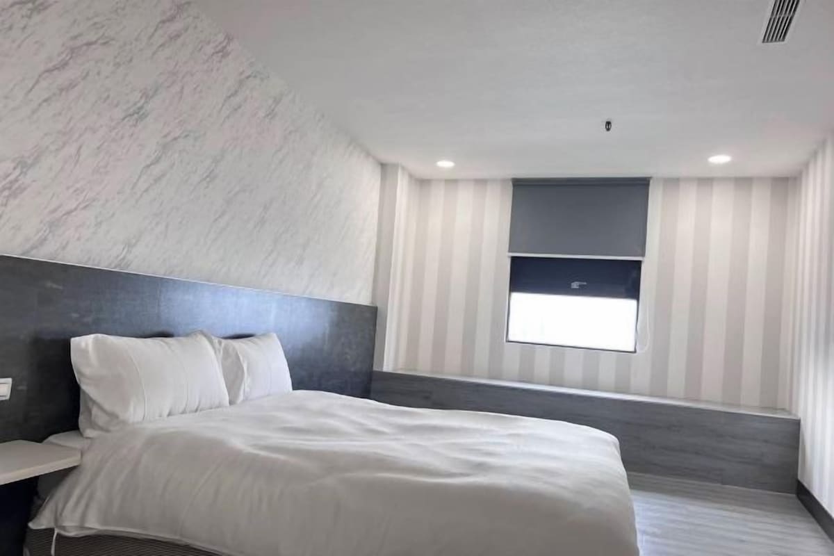 A modern hotel room with a large bed, a bedside table, a window with striped curtains, and a marble wall.