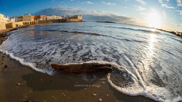 A beach with waves, a large rock, and a building with arches in the background.