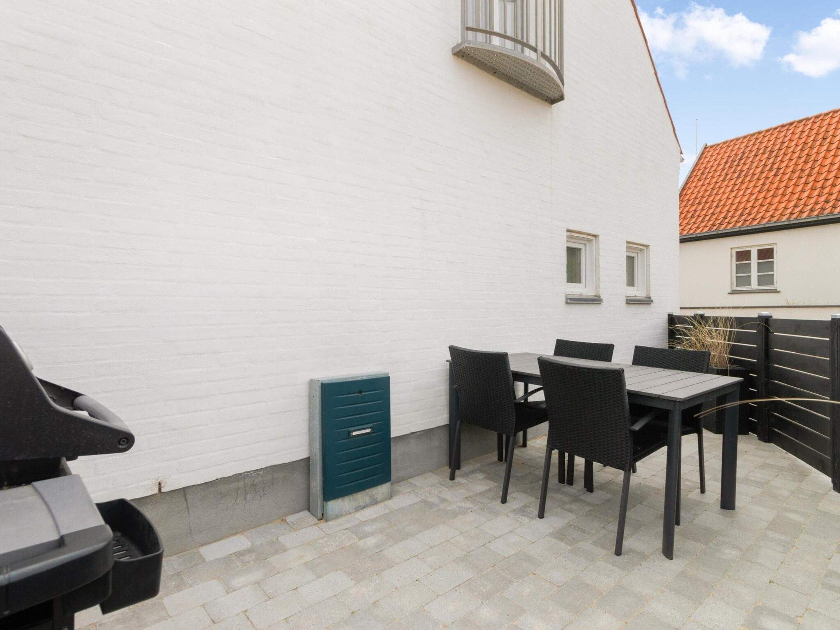 En terrasse med et bord og stole, en grøn el-tavle og en hvid bygning med rødt tegltag.
