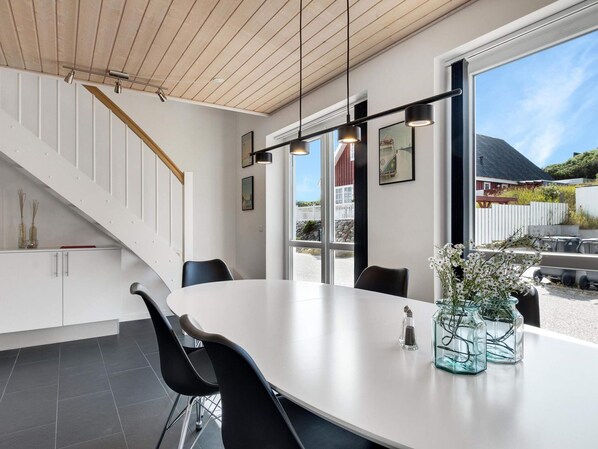 A modern interior with a white dining table, black chairs, a staircase, and large windows offering a view of the outside.
