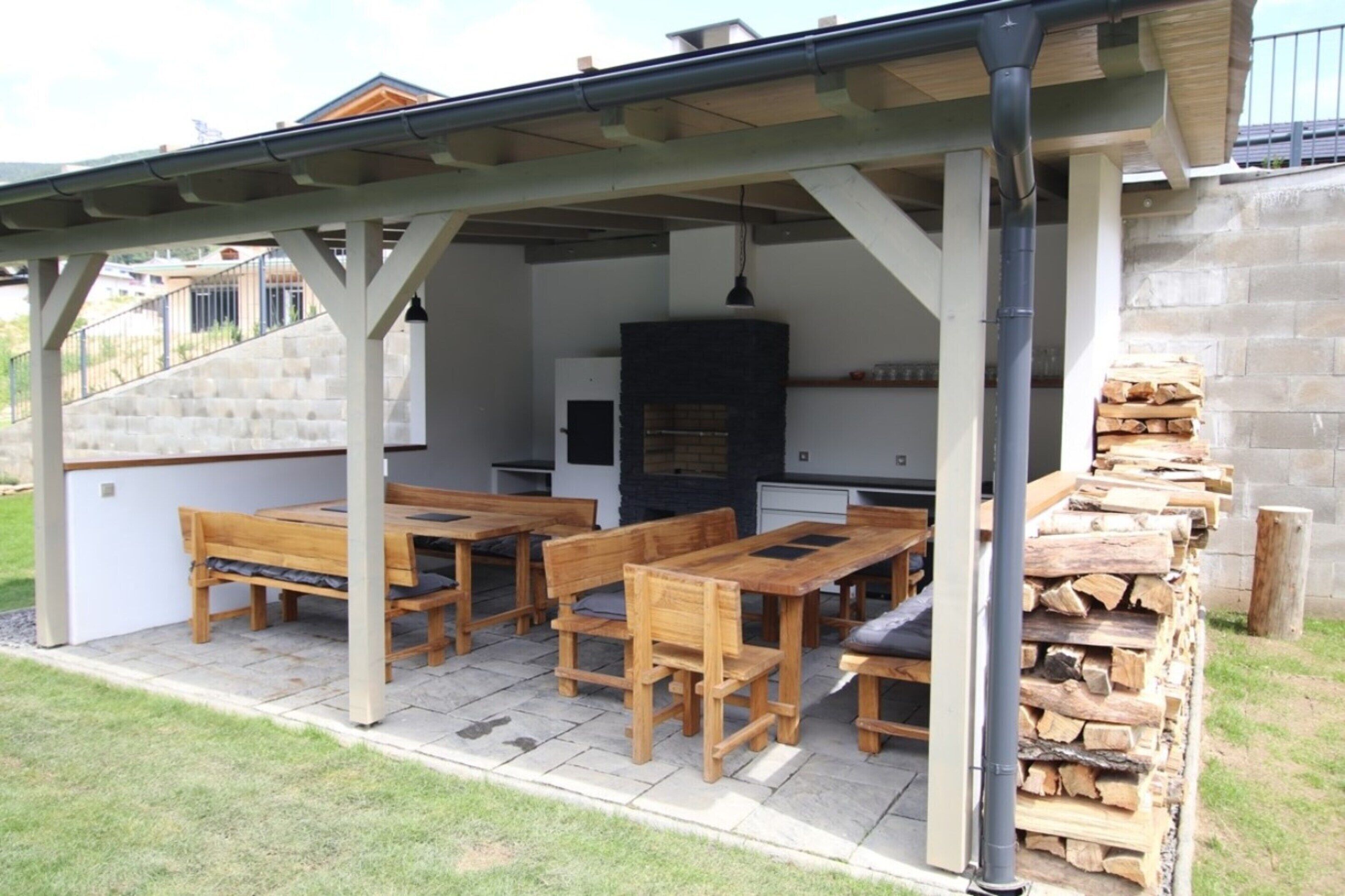 Een terras buiten met houten tafels en banken, een stenen open haard en een stapel haardhout.